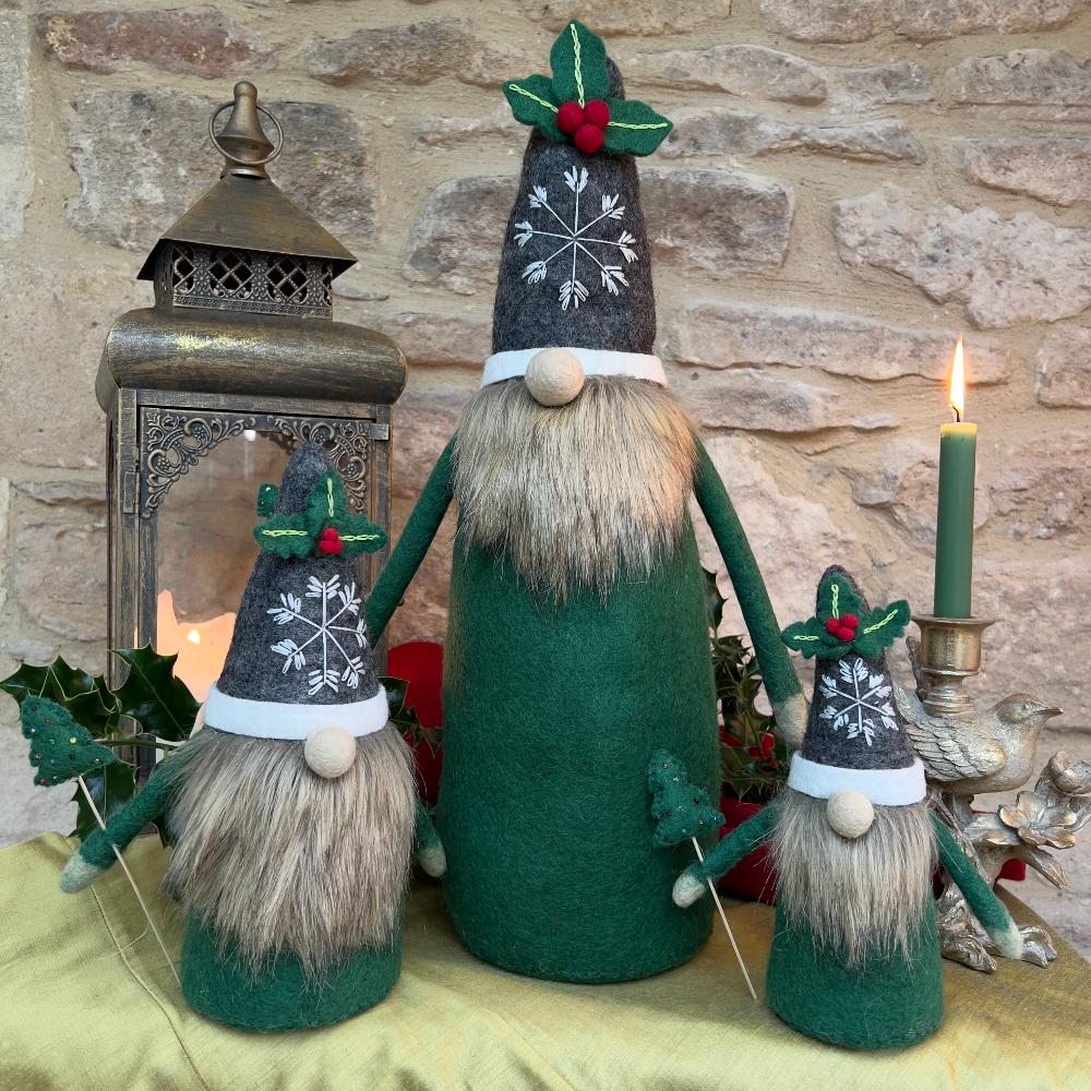 This felted wool gnome features a faux fur beard and wears a grey hat adorned with a hand-stitched snowflake and appliquéd holly with berries. He holds a removable Christmas tree embellished with hand-sewn beads. Shown against a stone wall background with a festive lantern and a lit candle. 