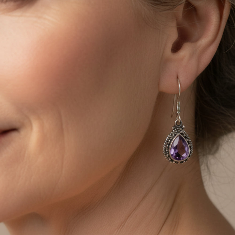 Amethyst & Silver Earrings against a cream wall background hanging from a branch with green leaves. 