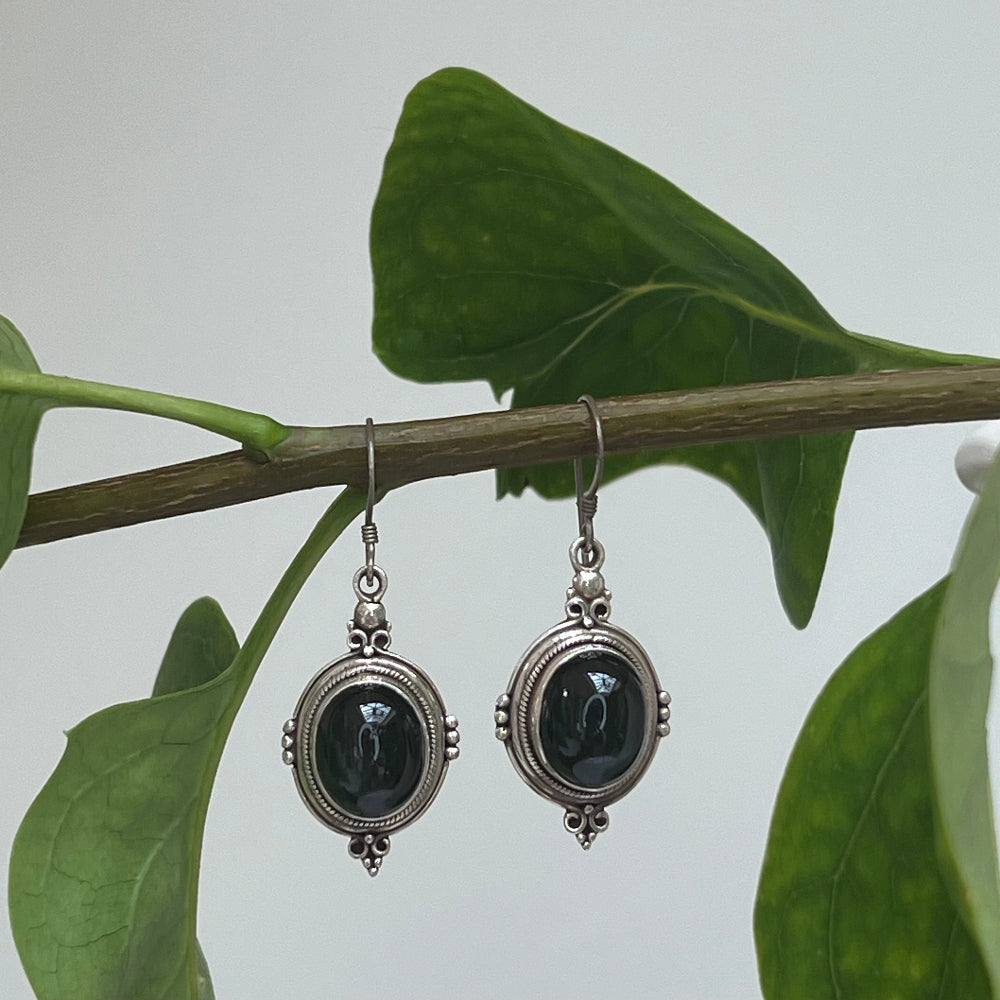 Black Star & Silver Earrings against a cream background hanging from  branch with green leaves. 