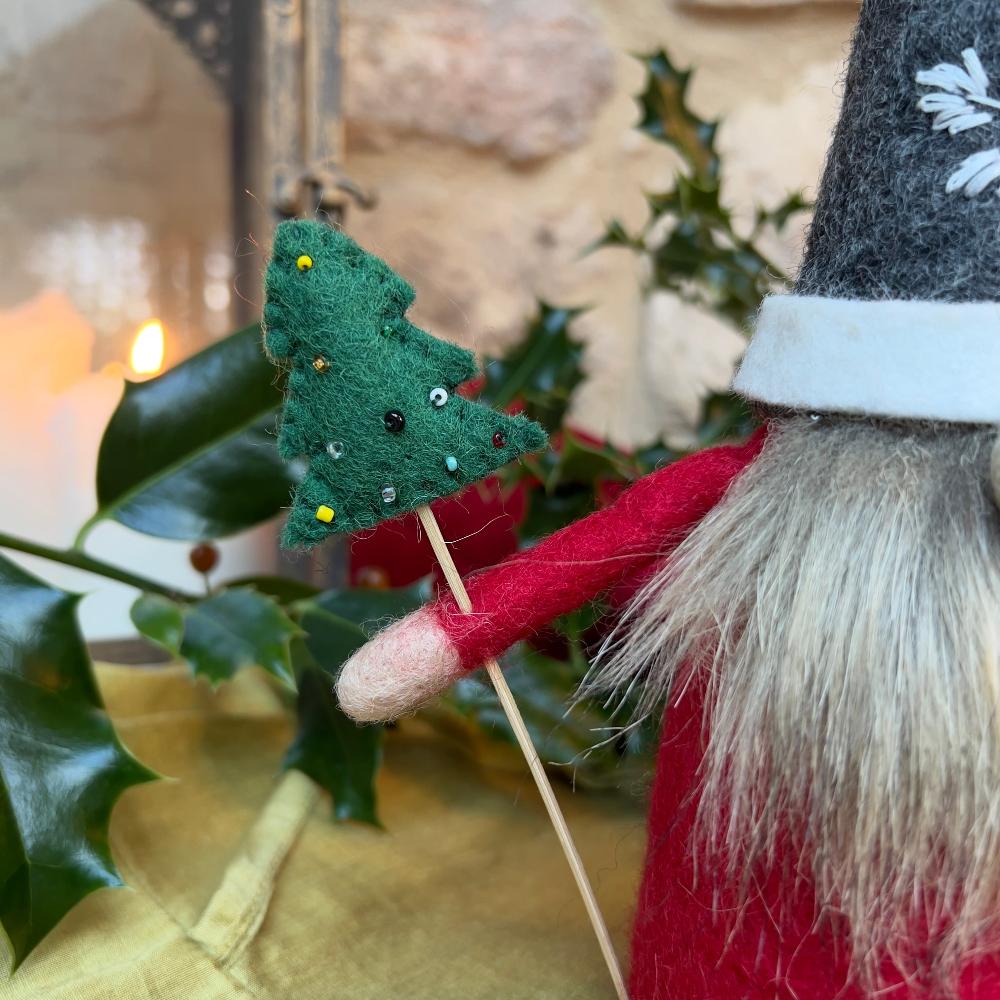 This felted wool gnome features a faux fur beard and wears a grey hat adorned with a hand-stitched snowflake and appliquéd holly with berries. He holds a removable Christmas tree embellished with hand-sewn beads. Shown against a stone wall background with holly and a festive lantern.
