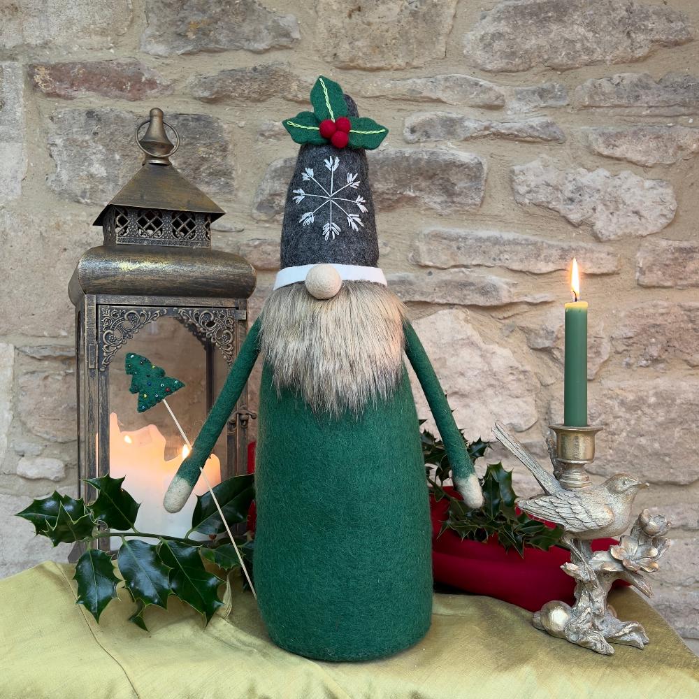 This felted wool gnome features a faux fur beard and wears a grey hat adorned with a hand-stitched snowflake and appliquéd holly with berries. He holds a removable Christmas tree embellished with hand-sewn beads.
