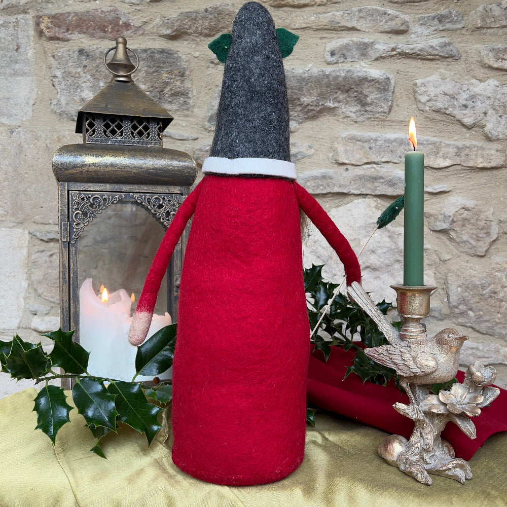 This felted wool gnome features a faux fur beard and wears a grey hat adorned with a hand-stitched snowflake and appliquéd holly with berries. He holds a removable Christmas tree embellished with hand-sewn beads. Shown against a stone wall background with a festive lantern and a bird ornament with green candle. 