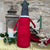 This felted wool gnome features a faux fur beard and wears a grey hat adorned with a hand-stitched snowflake and appliquéd holly with berries. He holds a removable Christmas tree embellished with hand-sewn beads. Shown against a stone wall background with a festive lantern and a bird ornament with green candle. 
