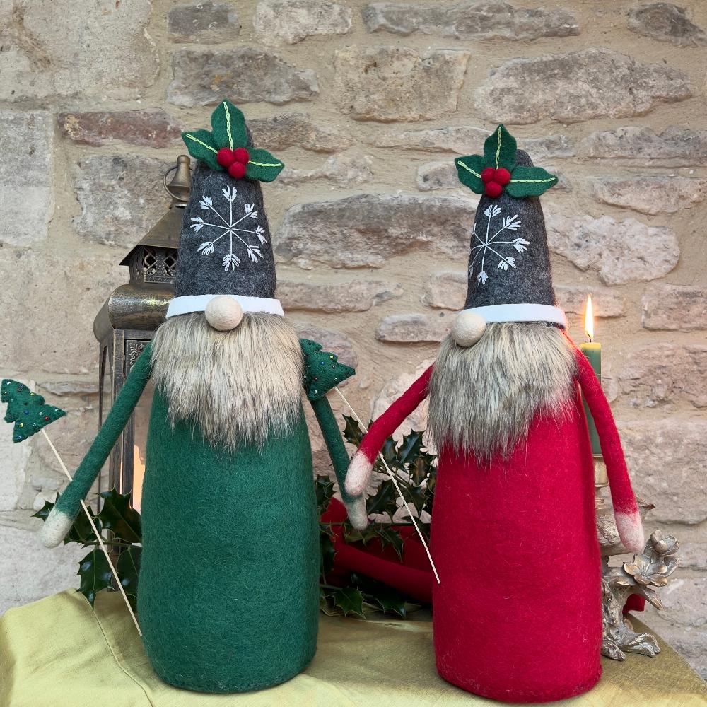 This felted wool gnome features a faux fur beard and wears a grey hat adorned with a hand-stitched snowflake and appliquéd holly with berries. He holds a removable Christmas tree embellished with hand-sewn beads.  Standing against a stone wall backdrop with green holly and a candle.