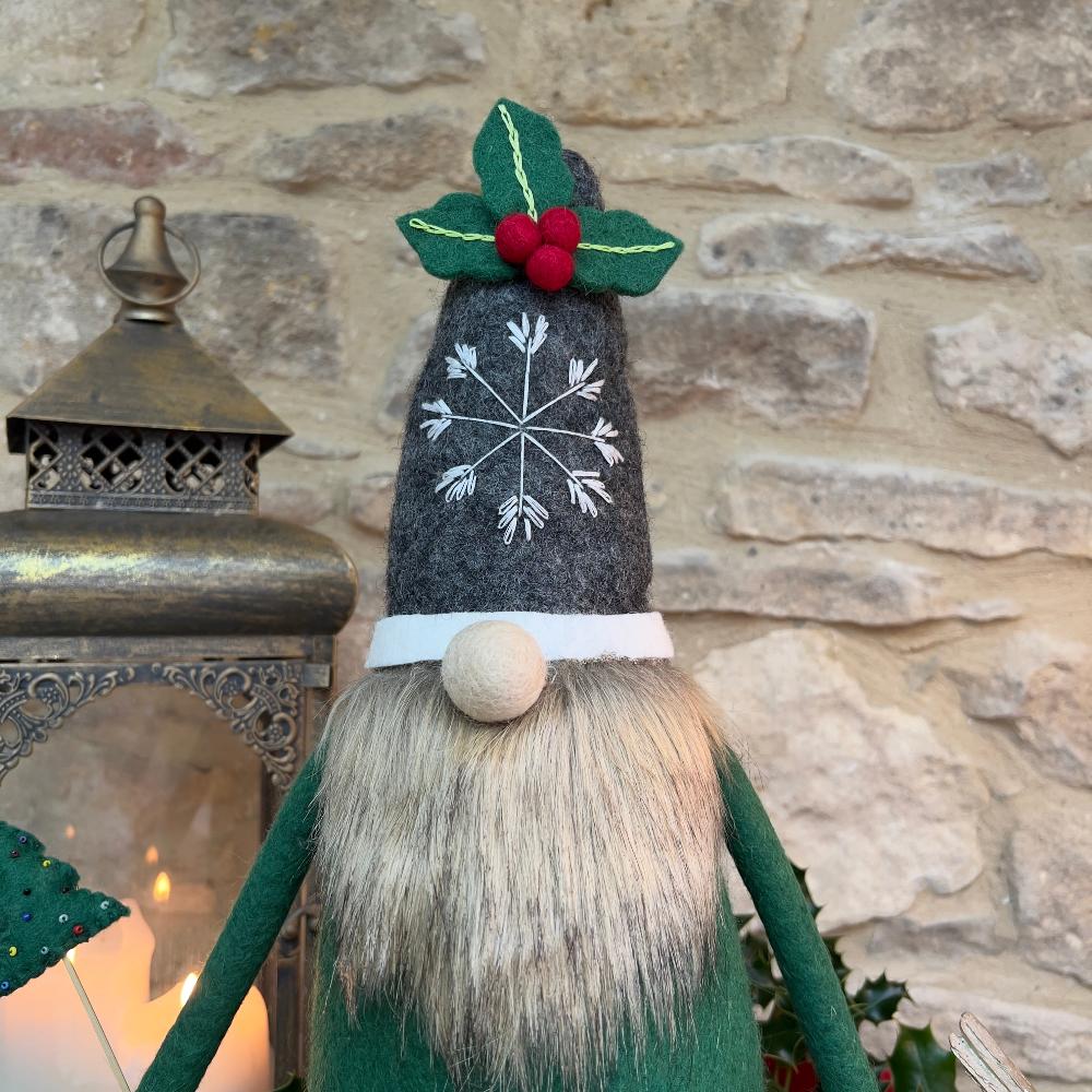 This felted wool gnome features a faux fur beard and wears a grey hat adorned with a hand-stitched snowflake and appliquéd holly with berries. He holds a removable Christmas tree embellished with hand-sewn beads.
