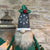This felted wool gnome features a faux fur beard and wears a grey hat adorned with a hand-stitched snowflake and appliquéd holly with berries. He holds a removable Christmas tree embellished with hand-sewn beads.