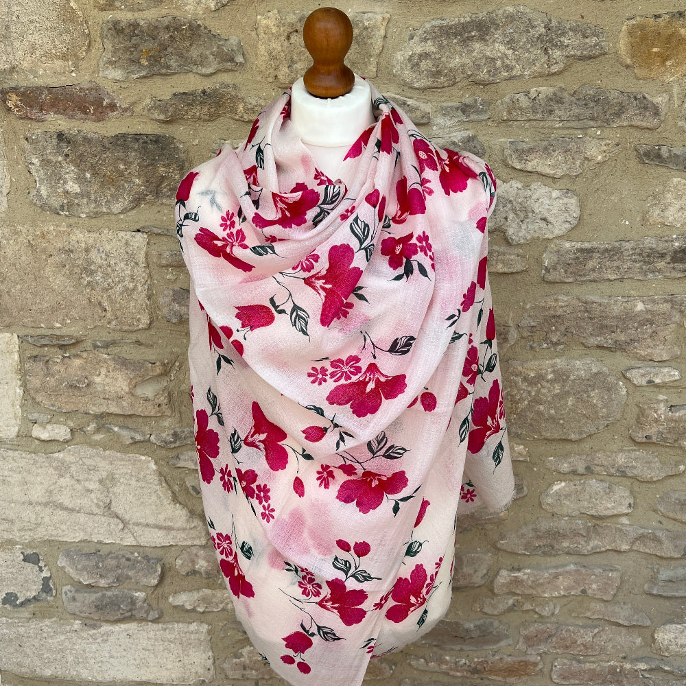 Floral cashmere scarf with pink flowers on a white background draped over a wooden mannequin against a stone wall.