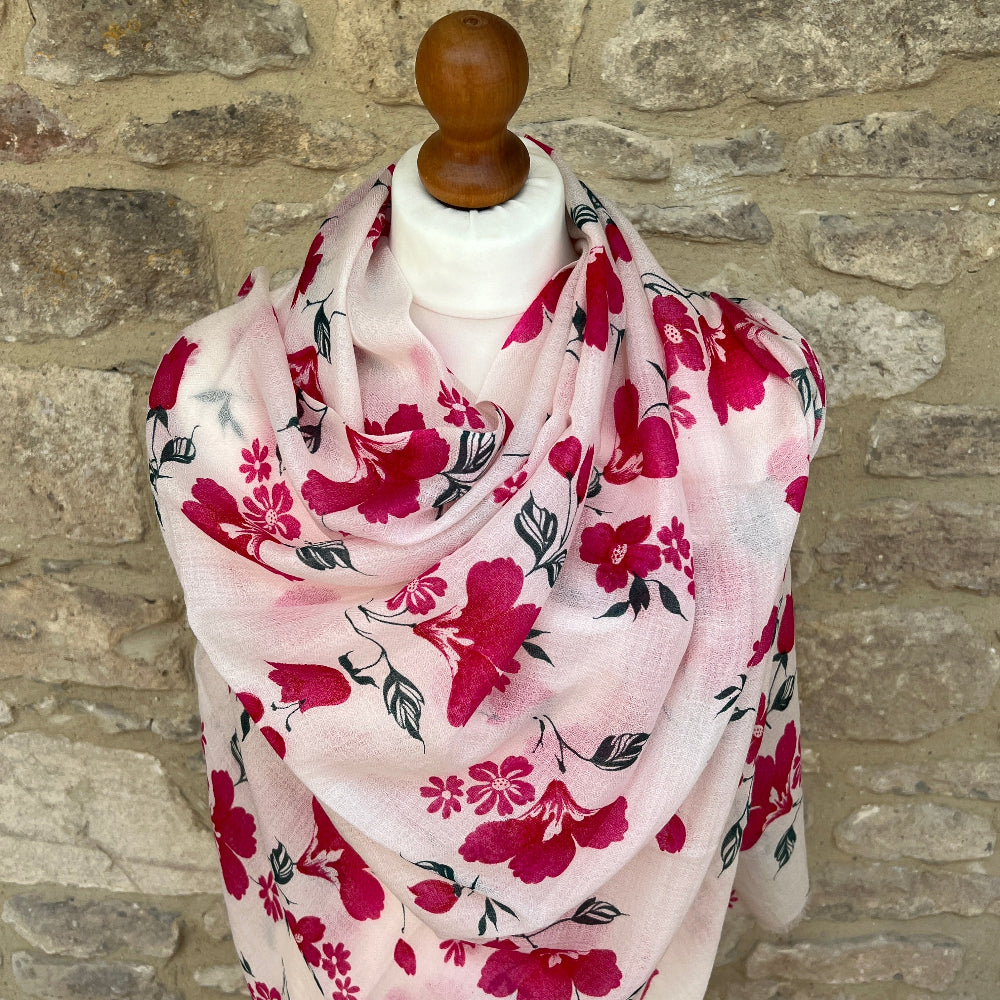 Floral cashmere scarf with pink flowers on a white background draped over a wooden mannequin against a stone wall.