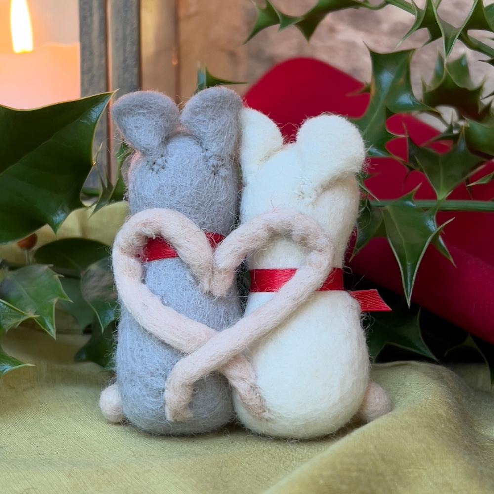 Introducing an enchanting duo of handcrafted felted wool mice, captured in a tender embrace this festive season.

Each mouse couple showcases hand embroidery and features poseable wired arms for display. Adorned with delicate red ribbon scarves and clutching sprigs of holly, these charming companions embody yuletide romance.

Their intertwined tails form a graceful heart—a poignant symbol of enduring affection. A refined addition to any Christmas collection, these artisanal pieces celebrate love 