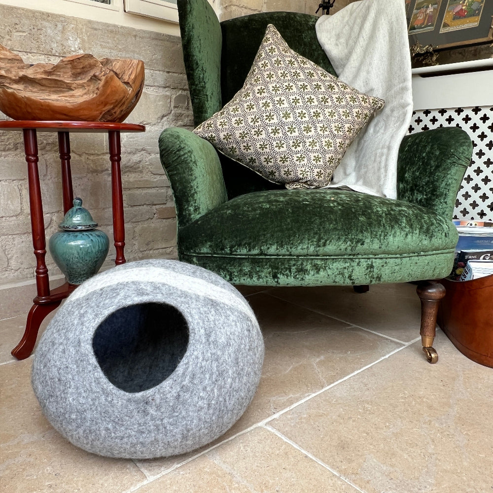 Gray pet bed  with a white stripe in front of a green velvet armchair with decorative pillows.
