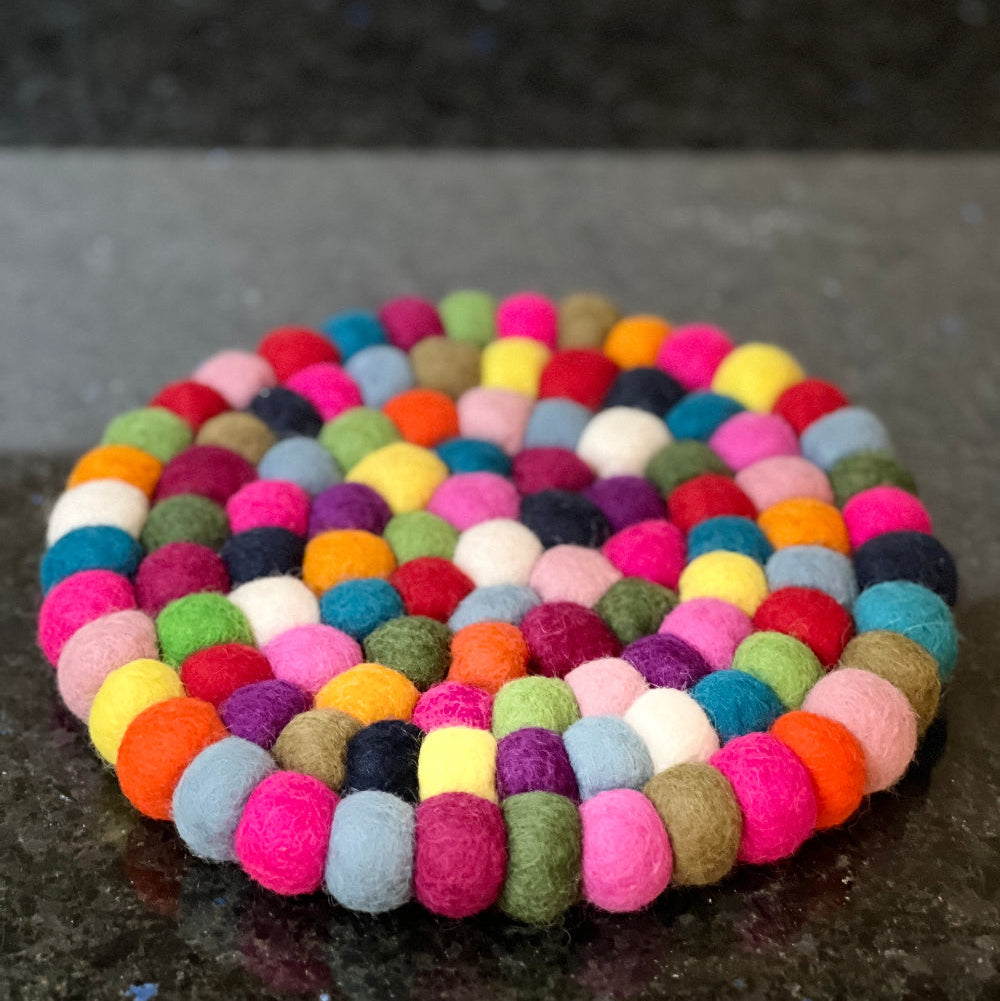 This handcrafted felted wool trivet combines vibrant multicoloured design with durable, sustainable materials.

Its hardwearing construction offers reliable protection for your surfaces while adding an elegant bright touch of colour to your home.

Shown with fresh fruit and a white pitcher jug on a granite work surface. 