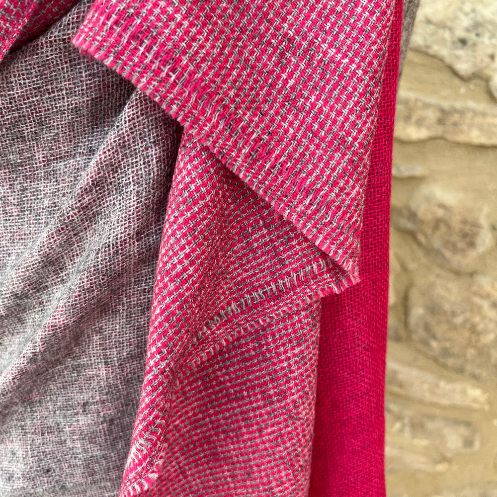 Handwoven Cashmere Reversible Pashmina - Pink &amp; Grey.  Mannequin  wearing a pink and gray cashmere scarf against a cotswold stone wall background
Close-up of a red and gray woven fabric with a textured background.