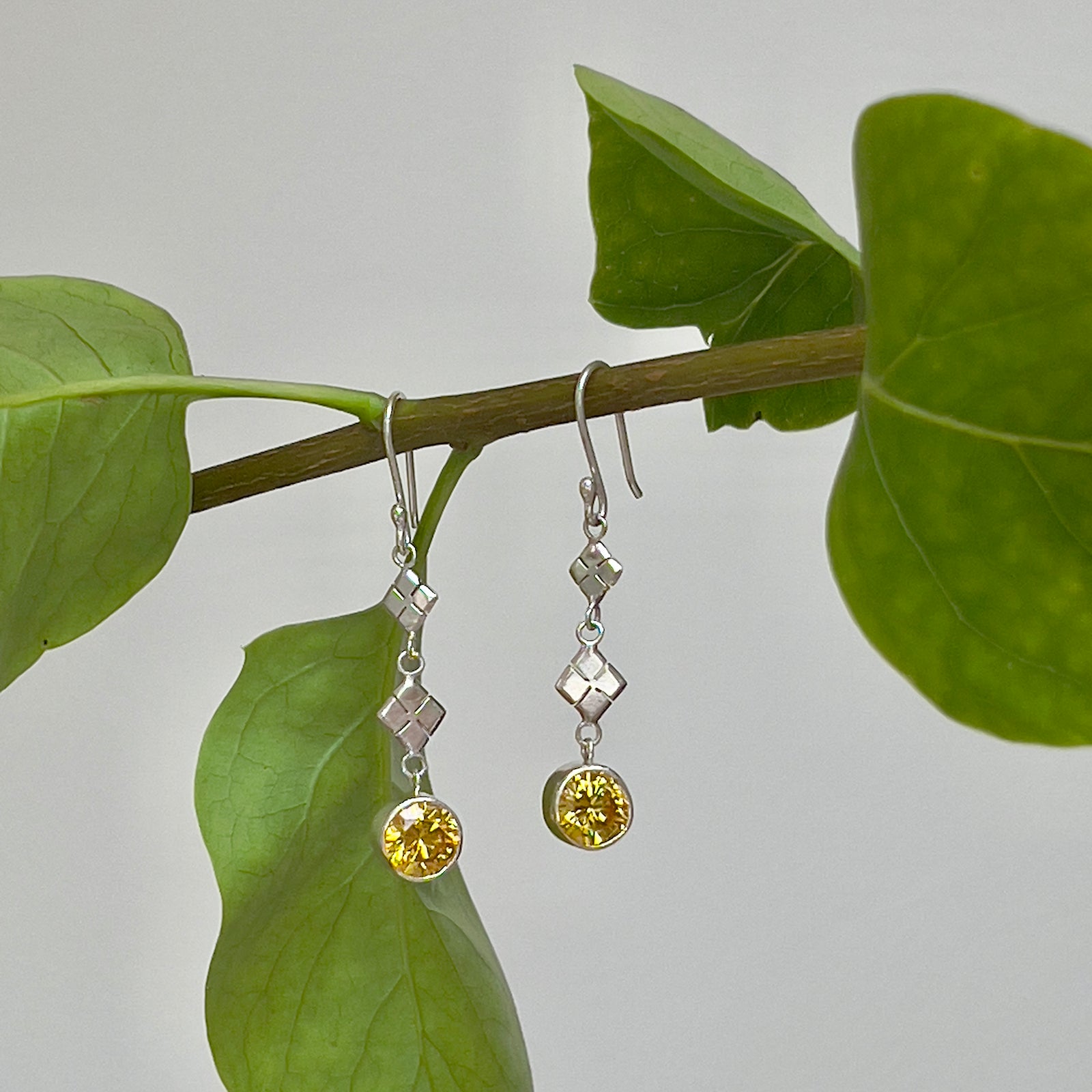 Yellow Topaz & Silver Drop Earrings against a cream wall background hanging from a branch with green leaves. 