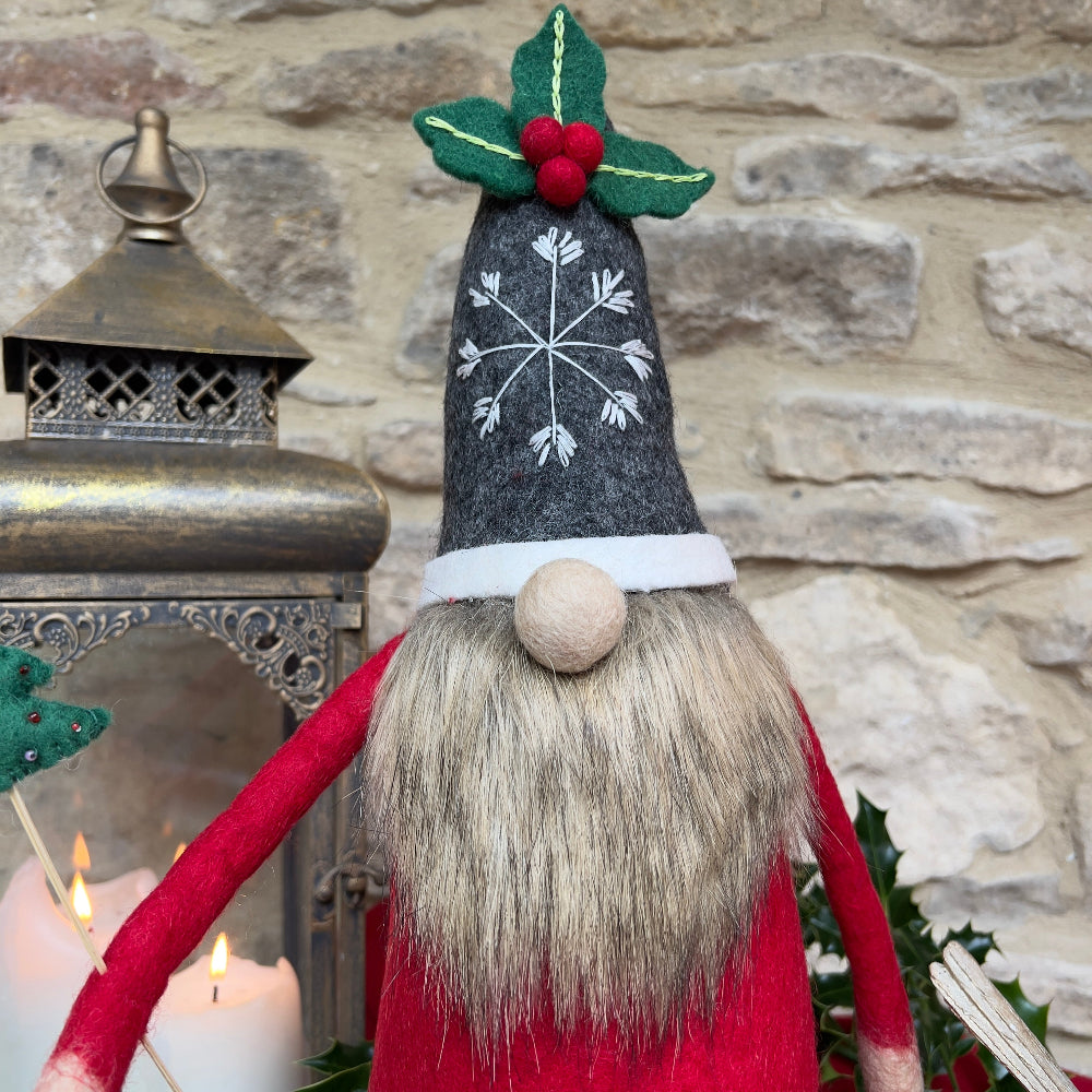 This felted wool gnome features a faux fur beard and wears a grey hat adorned with a hand-stitched snowflake and appliquéd holly with berries. He holds a removable Christmas tree embellished with hand-sewn beads. Shown against a stone wall background with a festive lantern and a bird ornament with green candle. 
