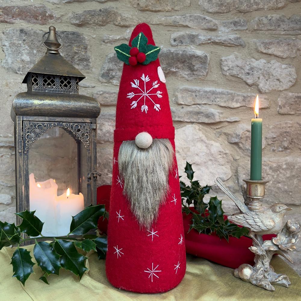 This Father Christmas gnome is crafted from felted wool, featuring a soft faux fur beard and delicately hand-stitched snowflake details on both hat and body. The hat showcases appliqué accents of holly, and berries, while the back is adorned with an exquisite appliquéd Christmas tree embellished with beads.