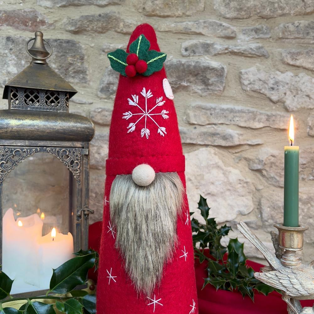 This Father Christmas gnome is crafted from felted wool, featuring a soft faux fur beard and delicately hand-stitched snowflake details on both hat and body. The hat showcases appliqué accents of holly, and berries, while the back is adorned with an exquisite appliquéd Christmas tree embellished with beads. Shown against a stone wall background, with a festive lantern and a candle. 