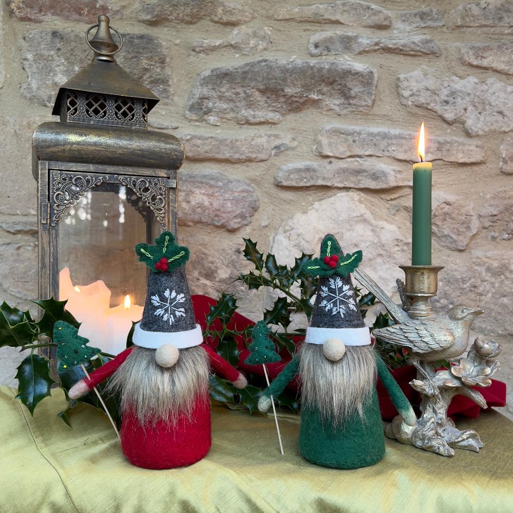 This felted wool gnome features a faux fur beard and wears a grey hat adorned with a hand-stitched snowflake and appliquéd holly with berries. He holds a removable Christmas tree embellished with hand-sewn beads. Shown against a stone wall background with holly and a festive lantern.