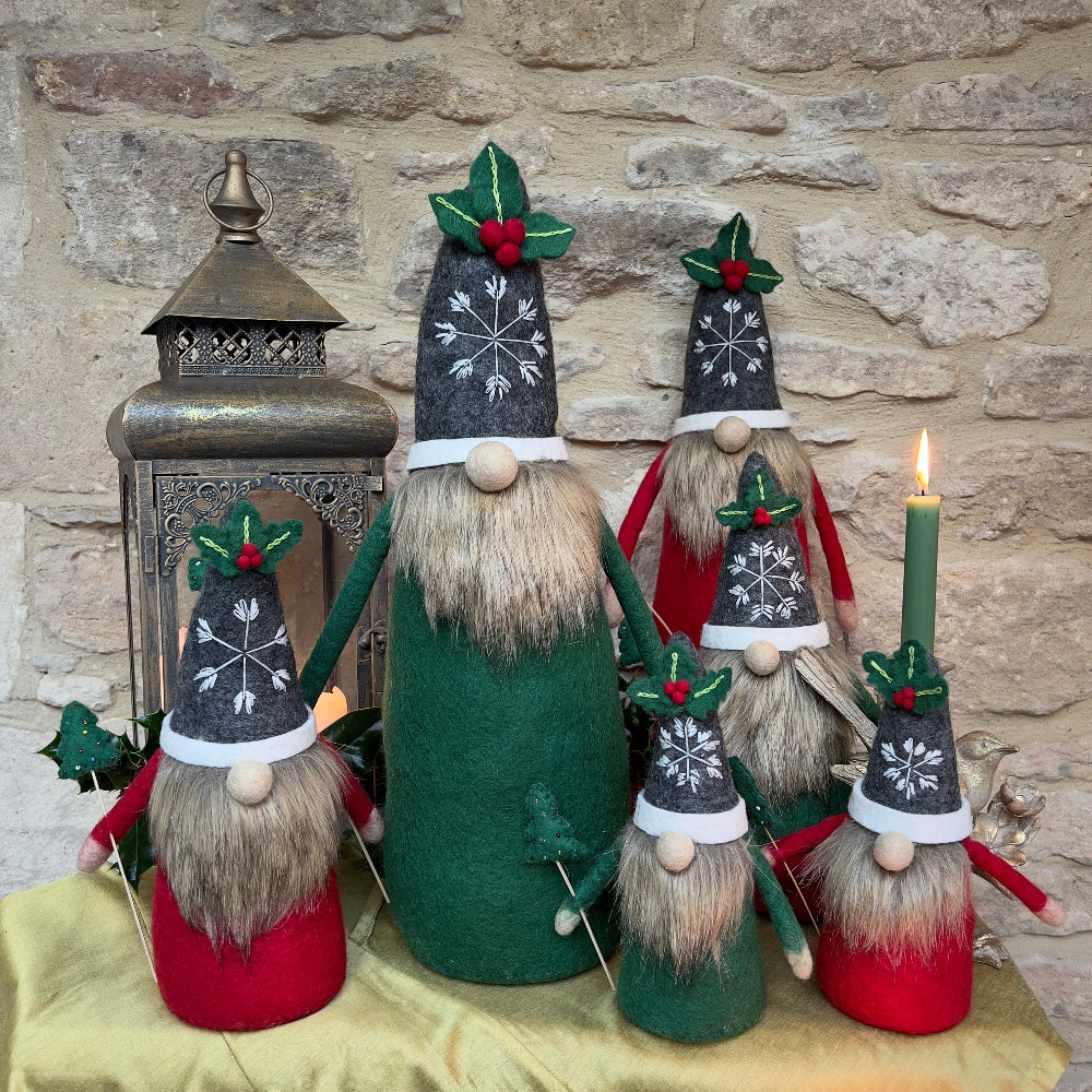 This felted wool gnome features a faux fur beard and wears a grey hat adorned with a hand-stitched snowflake and appliquéd holly with berries. He holds a removable Christmas tree embellished with hand-sewn beads. Shown against a stone wall background with a festive lantern and a bird ornament with green candle. 