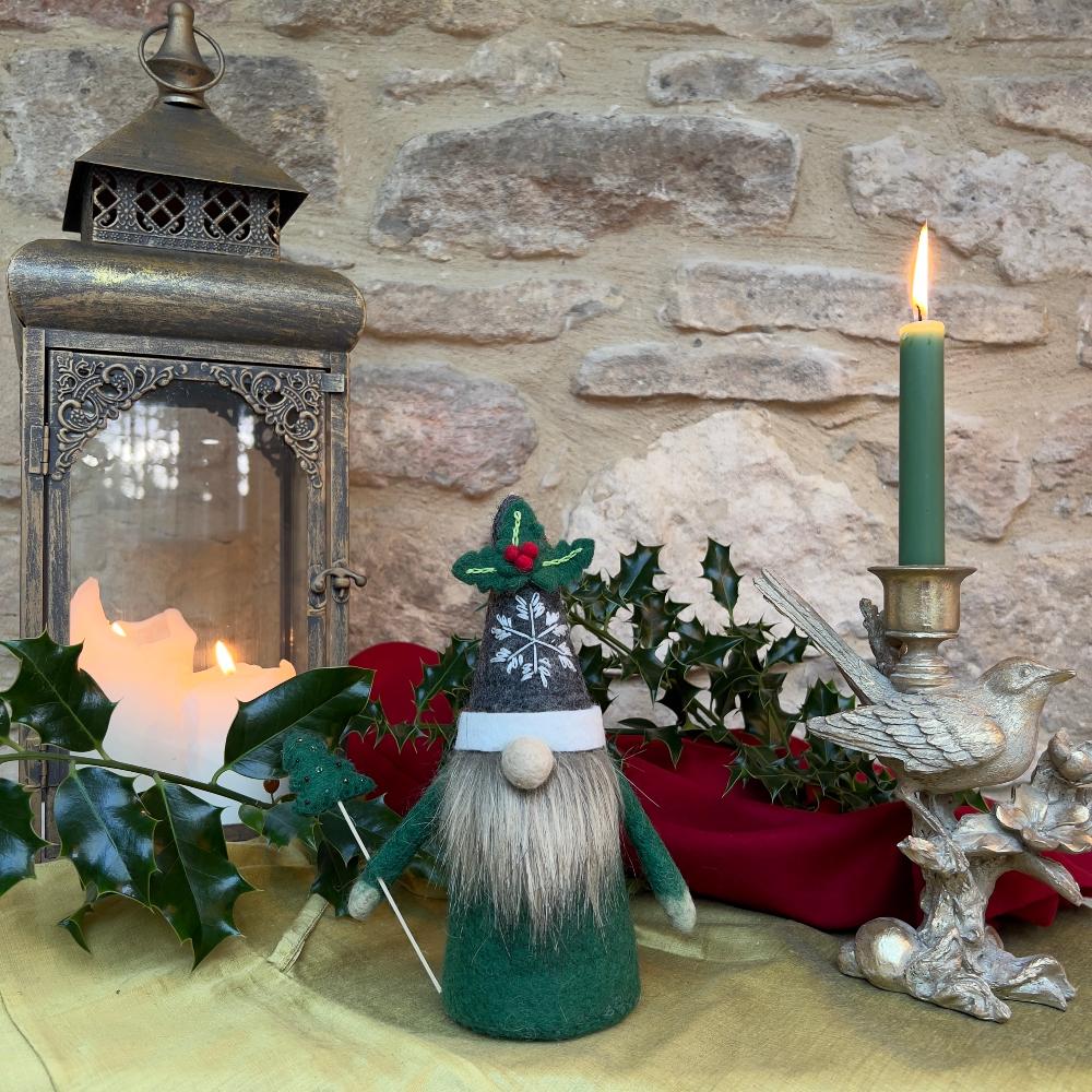 This felted wool gnome features a faux fur beard and wears a grey hat adorned with a hand-stitched snowflake and appliquéd holly with berries. He holds a removable Christmas tree embellished with hand-sewn beads.