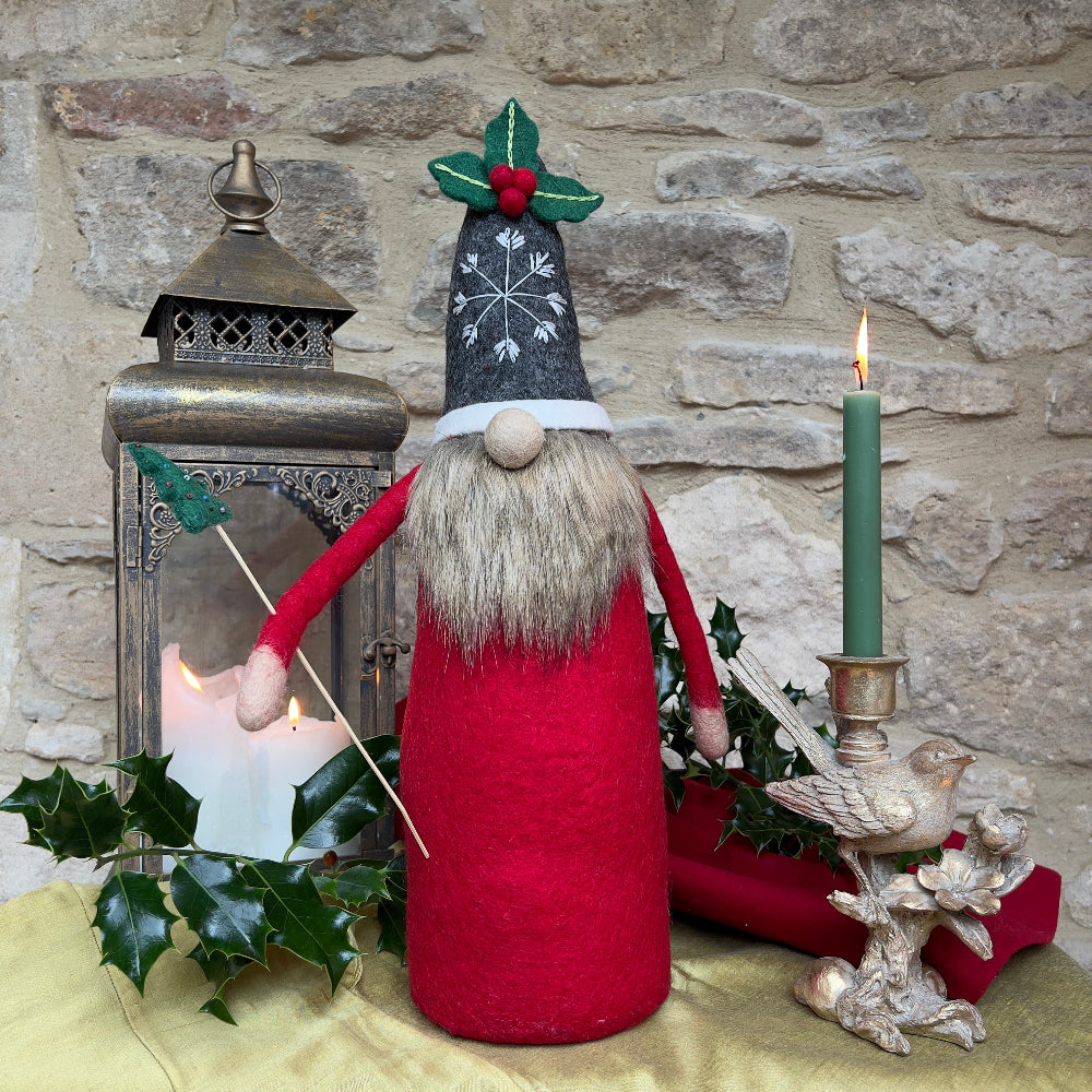 This felted wool gnome features a faux fur beard and wears a grey hat adorned with a hand-stitched snowflake and appliquéd holly with berries. He holds a removable Christmas tree embellished with hand-sewn beads. Shown against a stone wall background with a festive lantern and a bird ornament with green candle. 