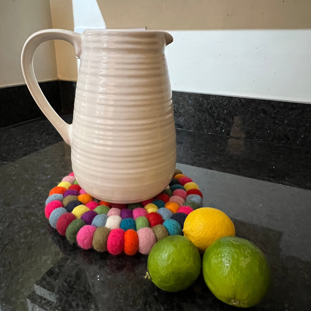 This handcrafted felted wool trivet combines vibrant multicoloured design with durable, sustainable materials.

Its hardwearing construction offers reliable protection for your surfaces while adding an elegant bright touch of colour to your home.

Shown with fresh fruit and a white pitcher jug on a granite work surface. 
