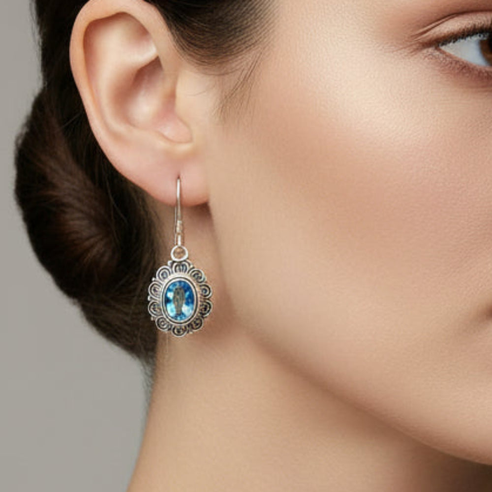 Topaz and silver earrings with an intricate design encasing the topaz stone hanging on a green leaved branch against a cream wall.