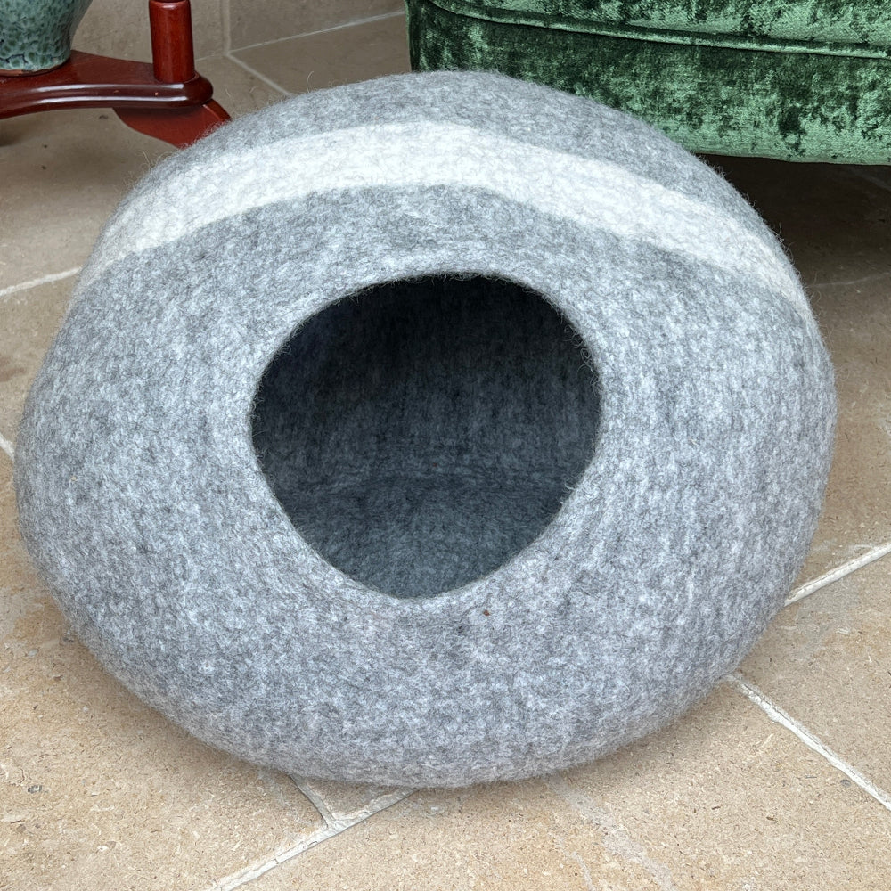Gray pet bed  with a white stripe in front of a green velvet armchair with decorative pillows.