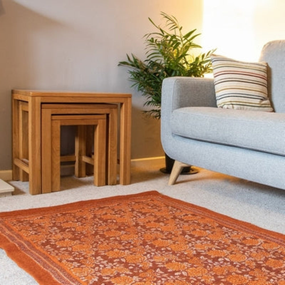 A burnt orange felted floral rug with a traditional rectangular shape, placed on a grey floor in a living room setting, next to a wooden stool and a grey sofa.