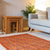 A burnt orange felted floral rug with a traditional rectangular shape, placed on a grey floor in a living room setting, next to a wooden stool and a grey sofa.