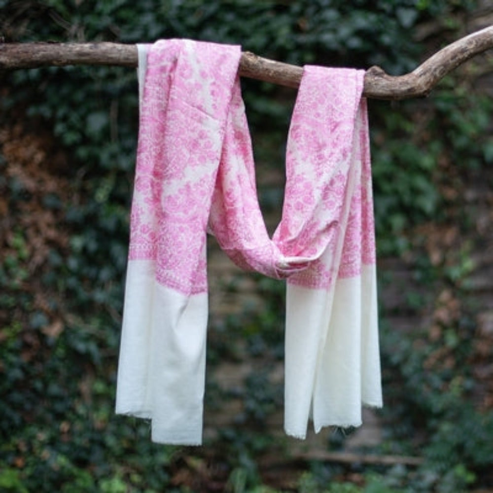 A cream and pink embroidered cashmere pashmina hanging on a branch of a tree with a green background.