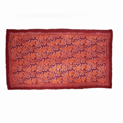 A handcrafted, rectangular, burgundy-colored felted wool rug by Douée with a floral pattern, placed on a neutral carpet with a grey sofa in a living room setting. A green plant and lamplight complete the room setting. 