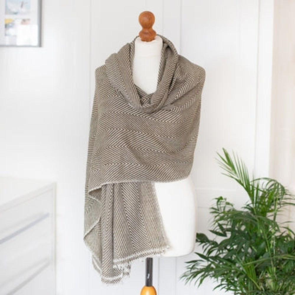 A sage green cashmere scarf with a herringbone pattern, displayed on a mannequin against a home background.