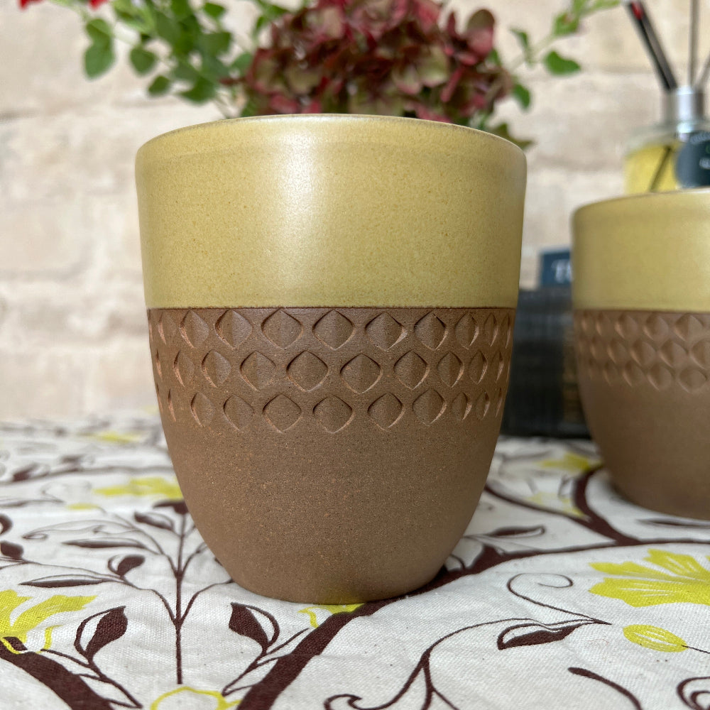 Hand Thrown and Decorated Mug - Bhaktapur Mustard colour on a patterned tablecloth, with coffee, books, room diffuser and fresh flower against a cotswold stone wall background. 