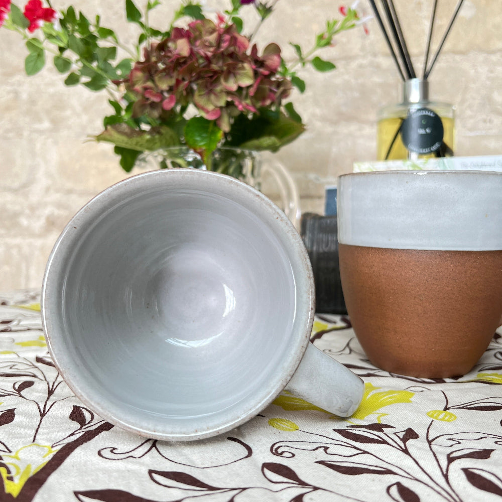 Hand Thrown Mug - Bhaktapur Silk on a patterned tablecloth with a scented room diffuser, fresh flowers shown against a cotswold stone wall