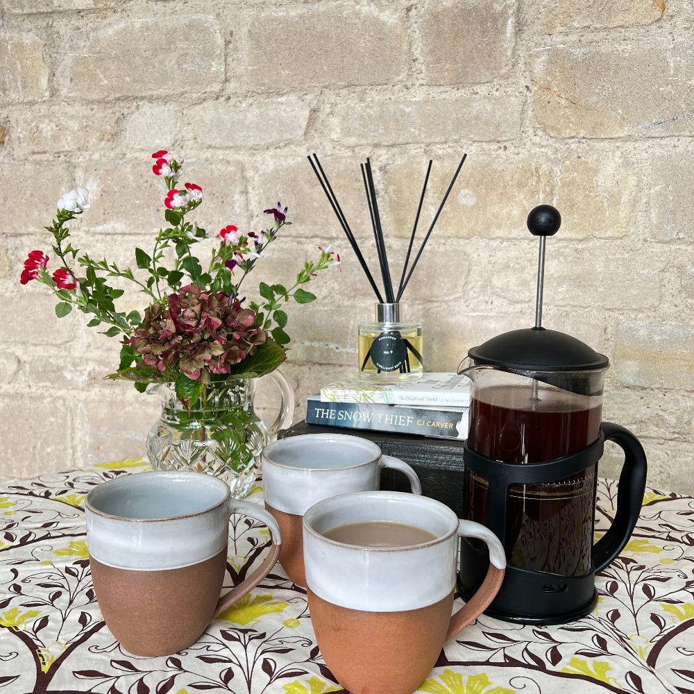 Hand Thrown Mug - Bhaktapur Silk on a patterned tablecloth with a scented room diffuser, fresh flowers and coffee.  Shown against a cotswold stone wall with the Snow Thief book by C J Carver