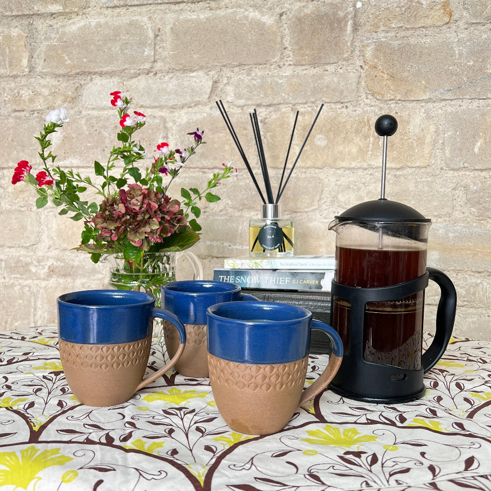 Hand Thrown and Decorated Mug - Bhaktapur Midnight Blue on a table with a Douée tablecloth, coffee, room diffusers and fresh flowers against a cotswold stone wall