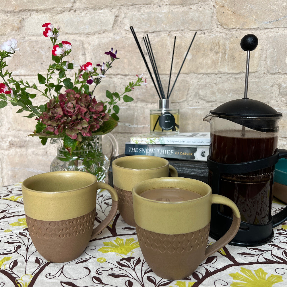 Hand Thrown and Decorated Mug - Bhaktapur Mustard colour on a patterned tablecloth, with coffee, books, room diffuser and fresh flower against a cotswold stone wall background. 