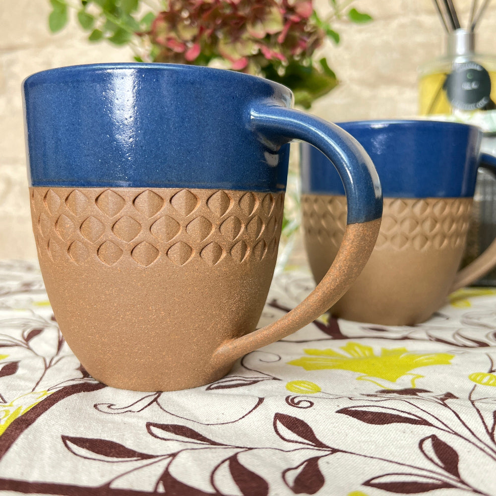 Hand Thrown and Decorated Mug - Bhaktapur Midnight Blue on a table with a Douée tablecloth, coffee, room diffusers and fresh flowers against a cotswold stone wall