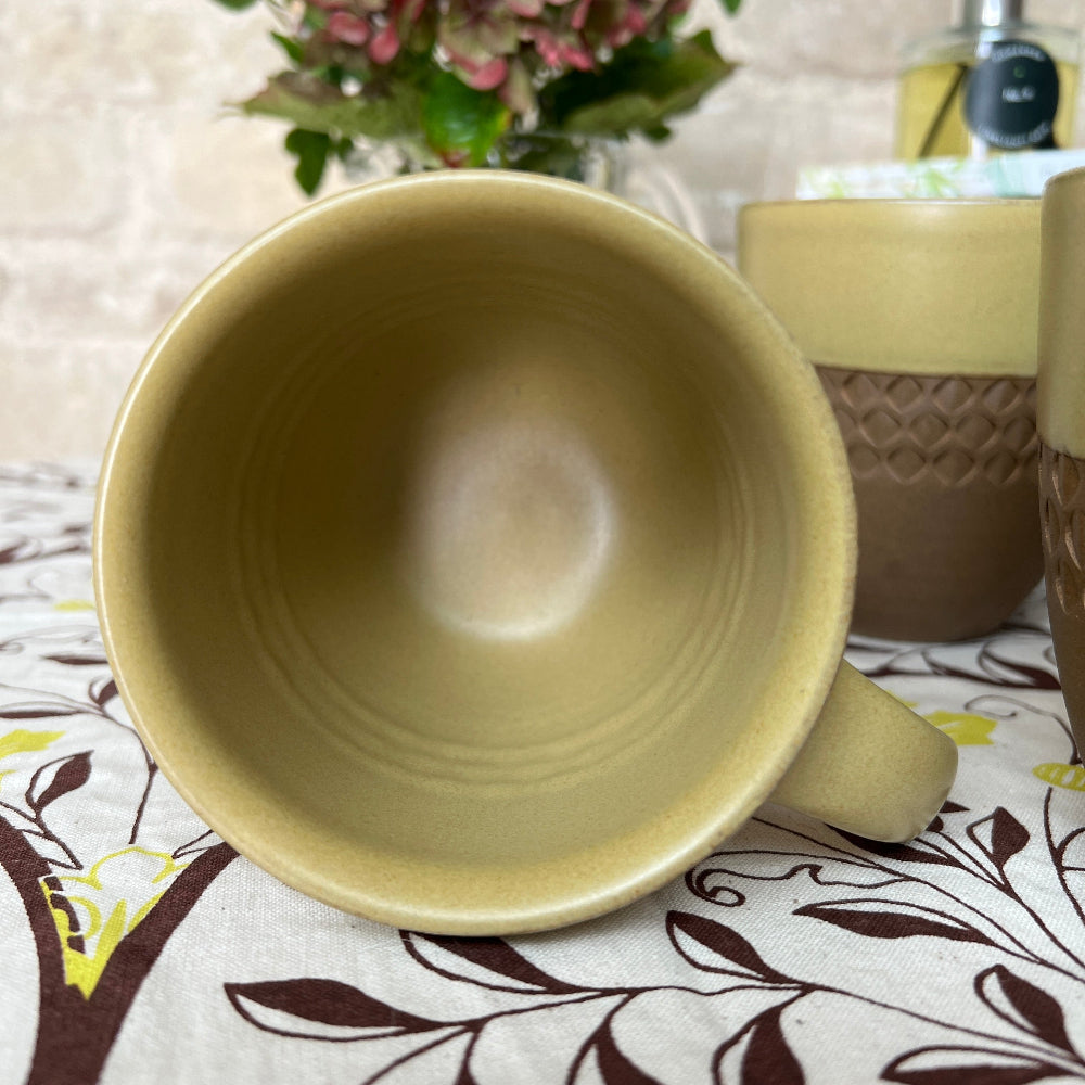 Hand Thrown and Decorated Mug - Bhaktapur Mustard colour on a patterned tablecloth, with coffee, books, room diffuser and fresh flower against a cotswold stone wall background. 