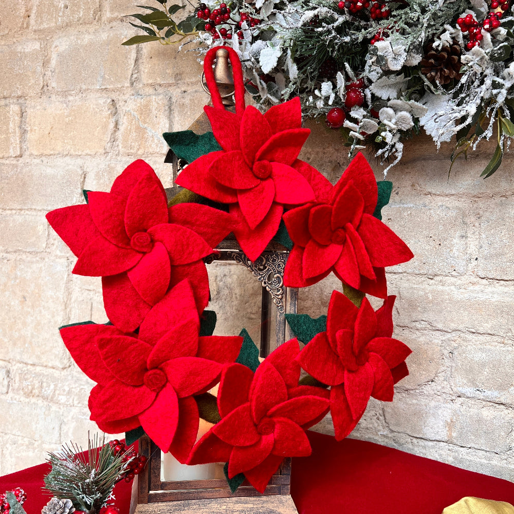 Handcrafted Felted Wool - &#39;Poinsettia&#39; Wreath with Red Poinsettia Flowers with Green leaves against a stone wool with christmas decor