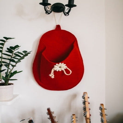 A red felted wool hanging storage slipper with a white appliqued flower and a wooden button centre, displayed on a wall.