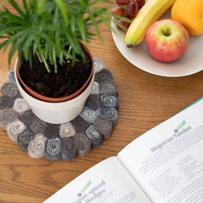 This handcrafted felted wool trivet combines a warm colour design with durable, sustainable materials. Shown with a green leafed plant, a plate of fresh fruit, a cookery book all on a wooden table. 