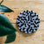 A round wool mat in black and grey colors, placed on a wooden surface with a green plant in the background.