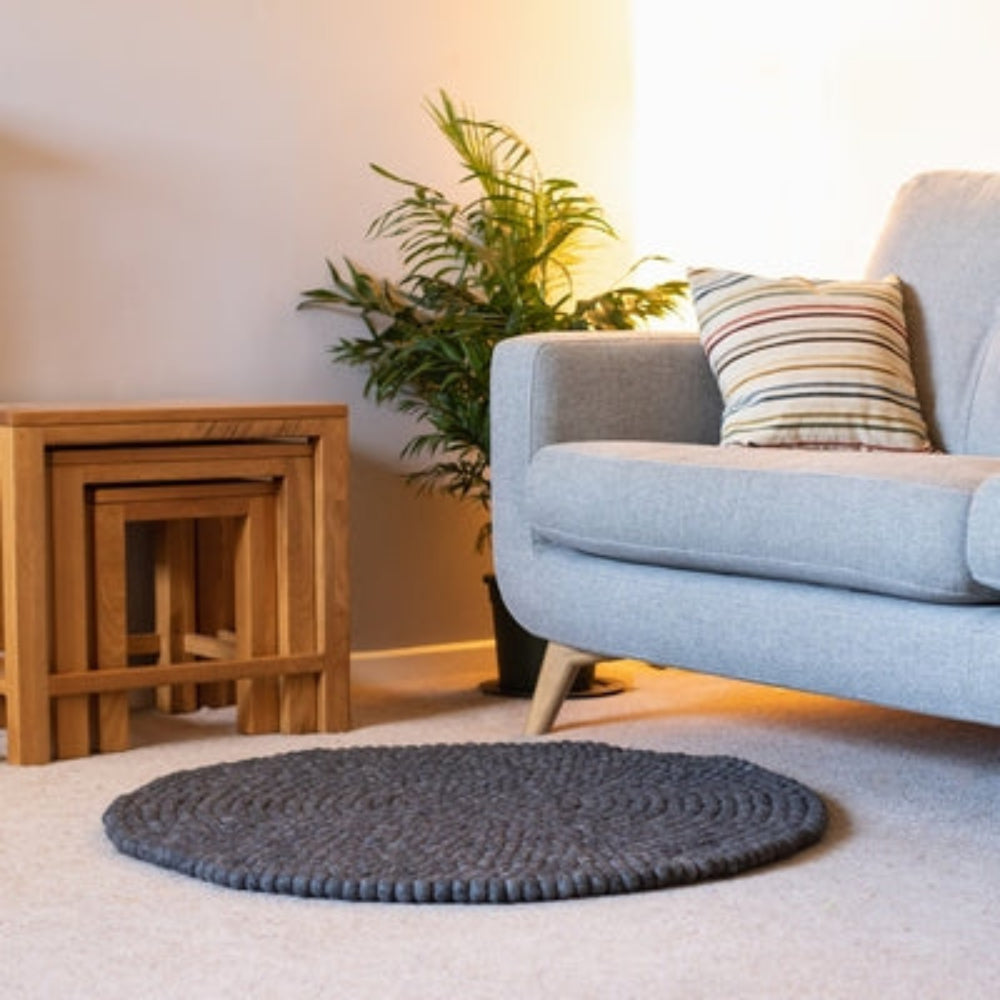 Handcrafted Felted Wool Rugs - Grey by Douée on a natural colour carpet in a room with tables and a natural colour sofa against a neutral wall with a green plant for decoration. 