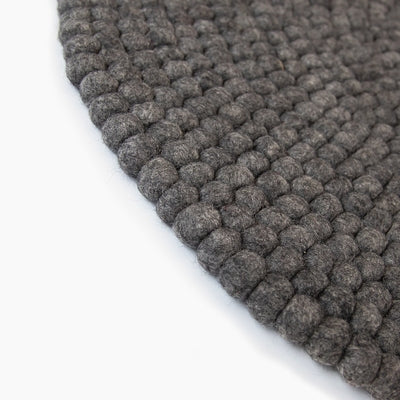 Handcrafted Felted Wool Rugs - Grey by Douée on a natural colour carpet in a room with tables and a natural colour sofa against a neutral wall with a green plant for decoration. 