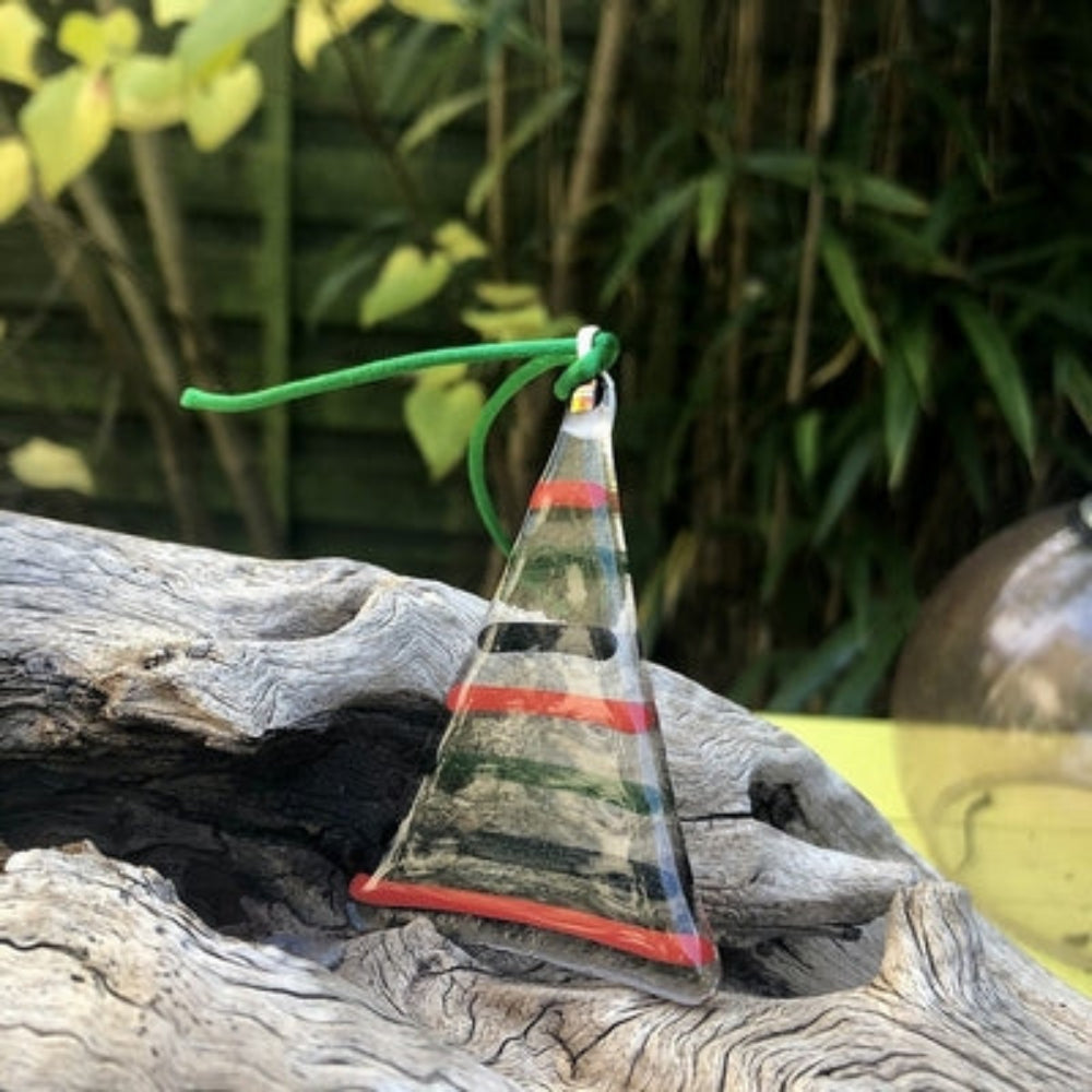 Handcrafted Glass Striped Christmas Tree with Red Ribbon shown against a wooden stand and green plants with grass. 