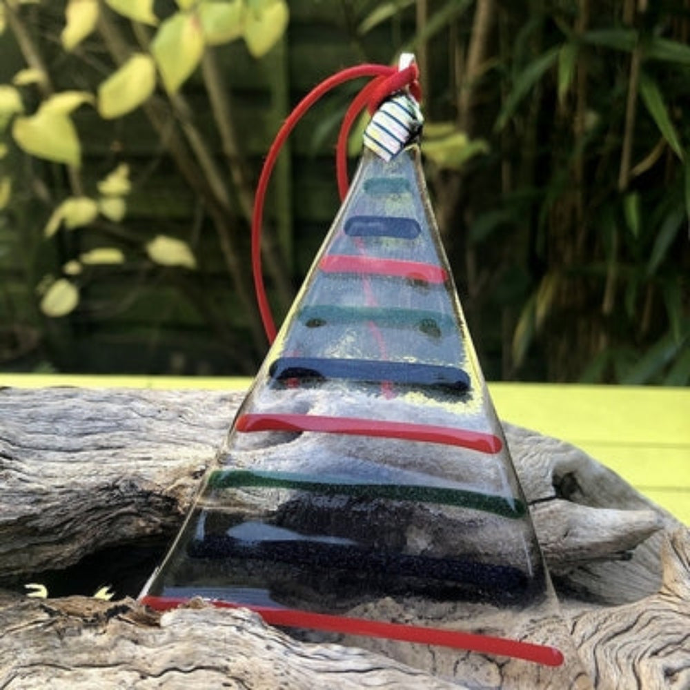 Handcrafted Glass Striped Christmas Tree with Red Ribbon shown against a wooden stand and green plants with grass. 