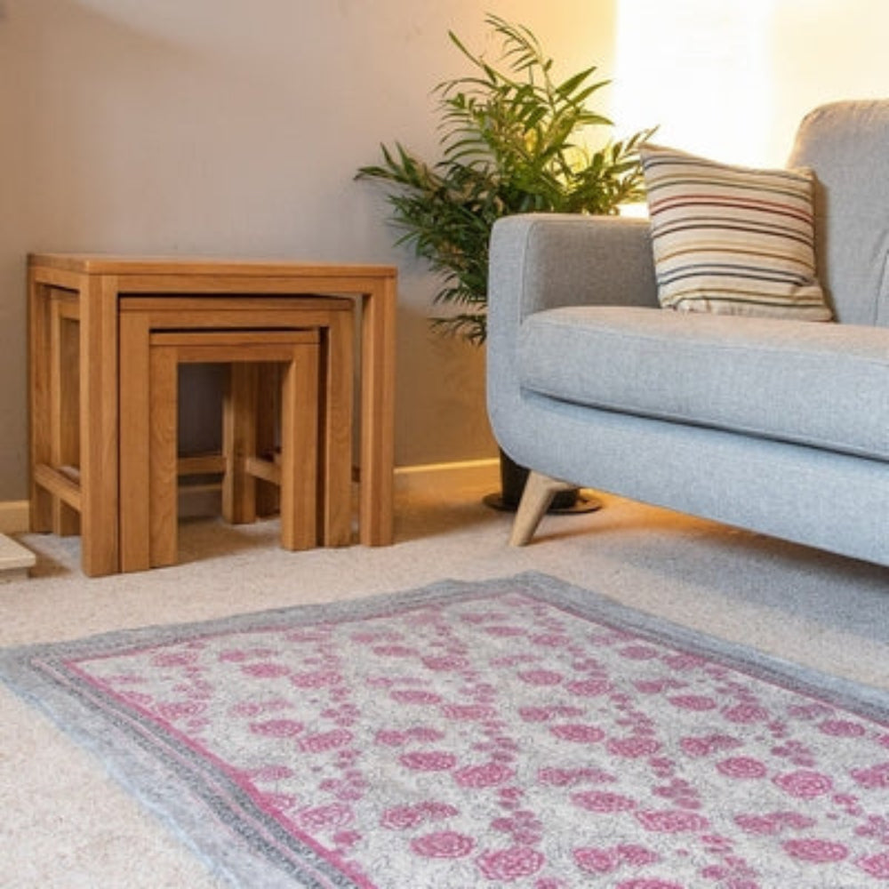 A gray floral rug with a patterned design, placed on a carpeted floor next to a wooden stool and a gray sofa, with a potted plant and a decorative cushion in the background.