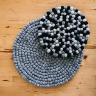 A round handcrafted sustainable wool trivet in black and grey colors, placed on a wooden surface with a green plant in the background.