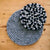 A round handcrafted sustainable wool trivet in black and grey colors, placed on a wooden surface with a green plant in the background.